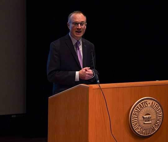 man with coat and tie, glasses, speaking at podium