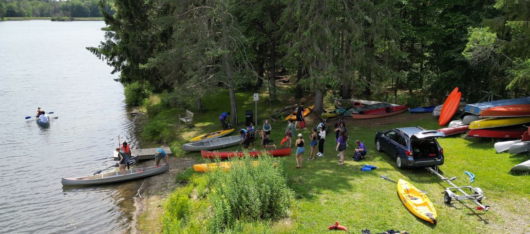 Students preparing canoes and kayaks at the shoreline of Foster Lake, with some people already paddling in the water and others gathered near parked vehicles and stacked boats under tall trees.