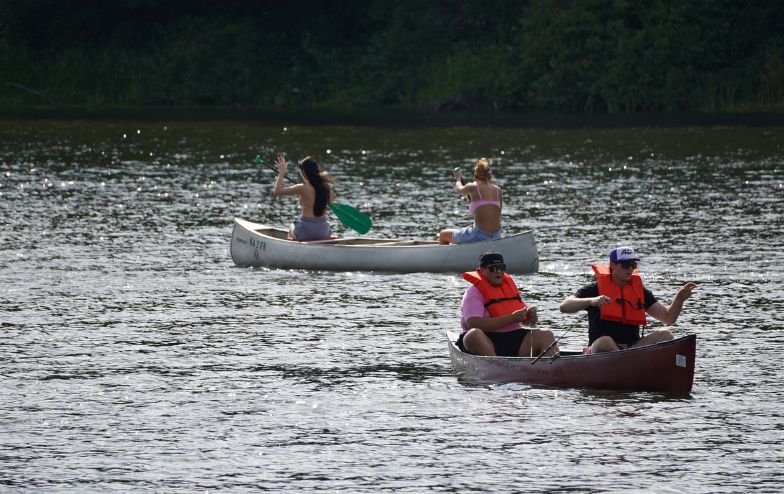 Two canoes on Foster Lake with students paddling; one canoe has two women in swimsuits, the other has two men wearing orange life vests and Alfred University hats, enjoying a sunny day on the water.