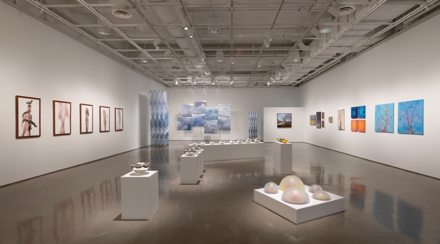 A spacious, modern art gallery with polished floors displaying a variety of student artworks, including paintings, sculptures, and mixed-media pieces arranged along the walls and on pedestals.