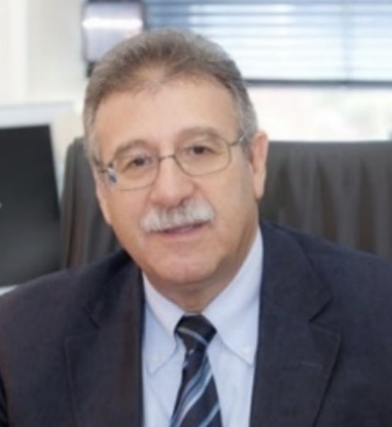 Man with gray hair and glasses wearing a suit and tie sits at a desk. He is smiling softly, giving the image a professional and approachable tone.