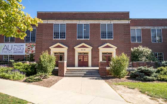 Exterior of a brick building labeled "Ade Hall" with three symmetrical doorways topped by triangular pediments, flanked by large windows.