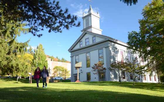 White building with a steeple and classical architecture, surrounded by green grass and autumn-colored trees under a clear blue sky. Two people in jackets walk across the lawn toward the entrance, suggesting a cool, sunny day on campus.