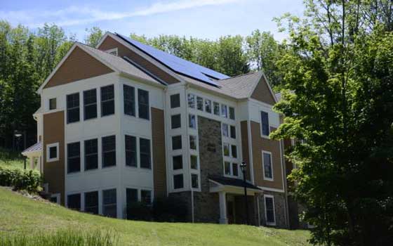 A modern, multi-story house with solar panels on the roof, nestled in lush greenery under a clear blue sky, conveys sustainability and tranquility.