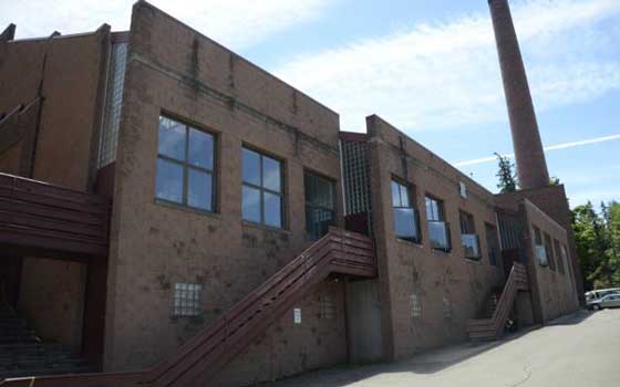 A large industrial brick building with multiple windows and two external staircases under a bright blue sky. A tall, narrow chimney is seen in the background.