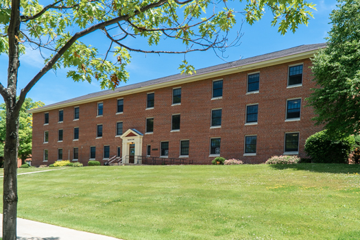 A red brick building with three stories and symmetrical windows stands amid green grass and trees under a clear blue sky, conveying a tranquil, academic setting.