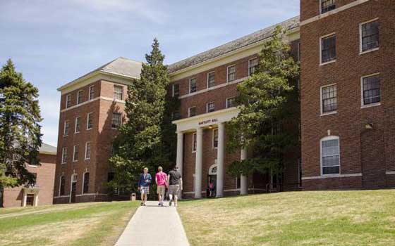 Three people walk towards a large brick building labeled "Bartlett Hall" surrounded by tall trees, on a sunny day, evoking a serene academic setting.