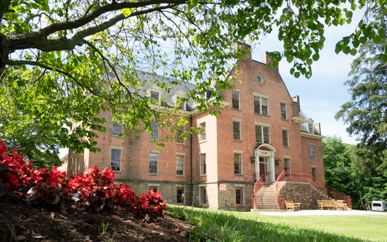 Historic brick building with arched windows and a central entrance, surrounded by lush greenery and red flowers. Sunny day, serene atmosphere.