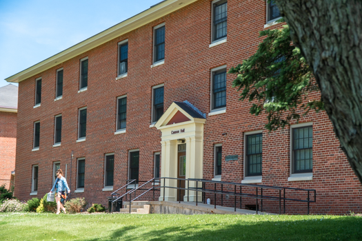 A person walks by a red brick building labeled "Cannon Hall," under a clear blue sky. The scene feels calm and serene, with green grass and trees.