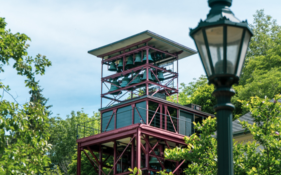A modern bell tower with a red metal frame stands among lush green trees under a blue sky. An antique-style streetlamp is visible in the foreground.