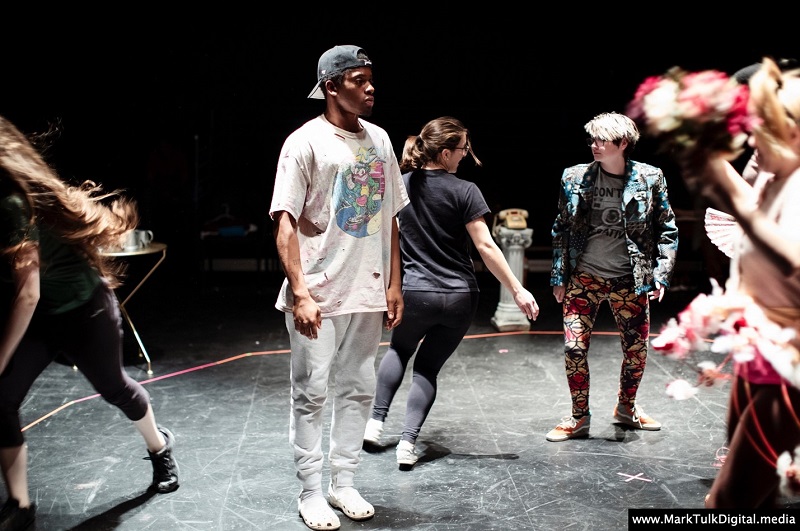 A rehearsal scene with diverse performers on stage, mid-movement. One wears a colorful t-shirt, another a patterned blazer. The mood is dynamic and focused.