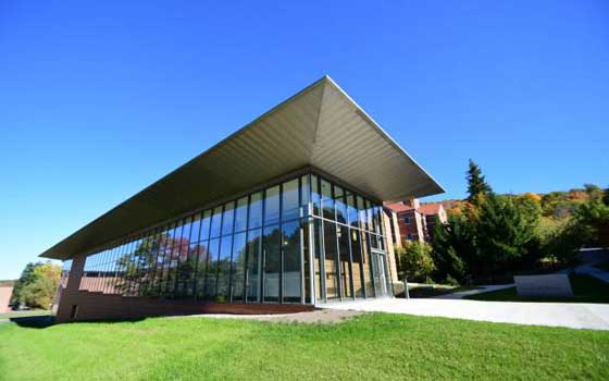 Modern building with a sharply angled roof and large glass facade, set on a grassy area with a sidewalk leading to the entrance. Autumn trees and brick buildings are visible in the background under a clear blue sky.