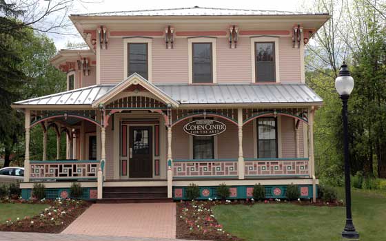 A two-story, Victorian-style house with a pink facade, elaborate trim, and a porch labeled "Cohen Center for the Arts." Surrounded by flowers and trees, it conveys a welcoming atmosphere.