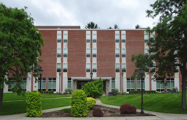 A four-story red-brick building with large windows, framed by green trees and shrubs. Overcast sky above, creating a calm and tranquil atmosphere.