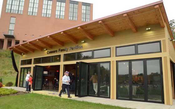 Single-story building with wooden roof and glass doors labeled "Connors Family Pavilion." People entering, grass in foreground, brick building behind.