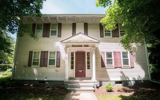 A two-story building labeled "Crandall Hall" with white siding, red shutters, and a red door. Surrounded by lush greenery on a sunny day.