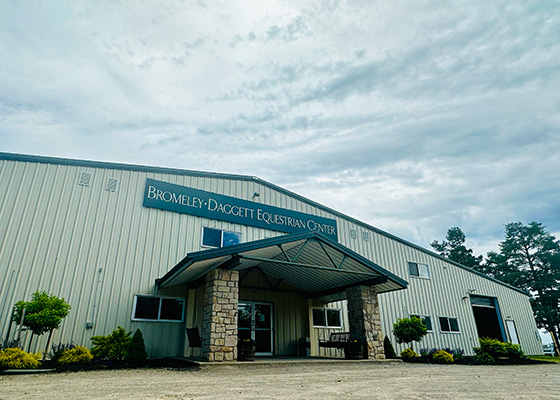 Large metal building with stone columns at the entrance, labeled "Bromley-Daggett Equestrian Center." Overcast sky and greenery surround the structure, conveying a calm and open atmosphere.