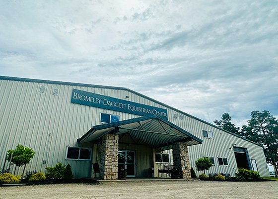 Large metal building with stone columns at the entrance, labeled "Bromley-Daggett Equestrian Center." Overcast sky and greenery surround the structure, conveying a calm and open atmosphere.
