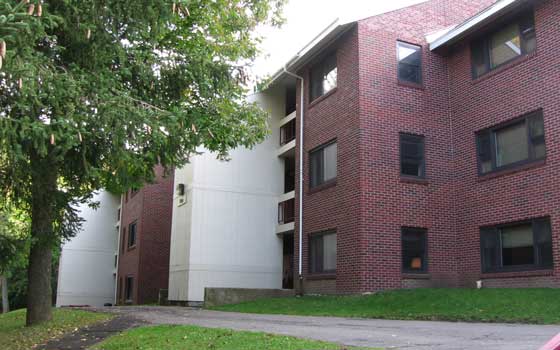 Red brick apartment building with three stories, surrounded by green grass and a tree to the left. The setting is calm and suburban.