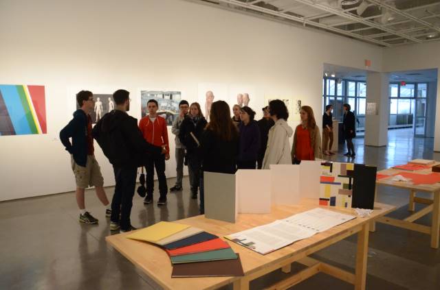 Group of people in an art gallery with modern art on the walls. A table displays colorful papers and materials. Bright, open space with a conversational tone.