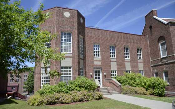 Red-brick building with large windows, surrounded by green shrubs and a tree, set under a clear blue sky. The scene feels welcoming and serene.