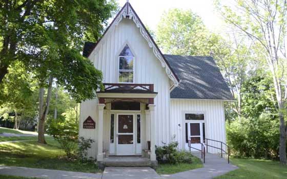 A charming, white, gabled cottage with ornate trim sits amid lush green trees under bright sunlight. Paths lead to the entrance.