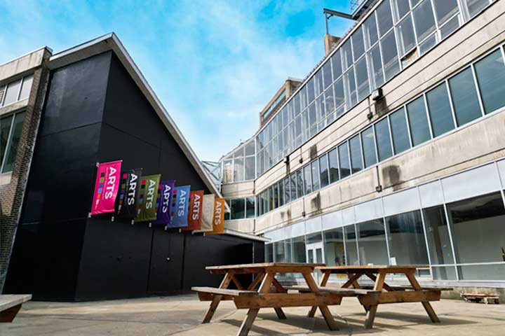 Exterior of a modern arts building with colorful "Arts" banners. The courtyard has wooden picnic tables on a sunny day, creating an inviting atmosphere.