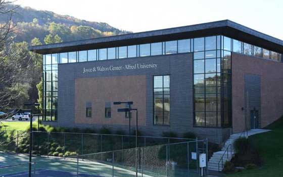 Modern building with large windows labeled "Joyce & Walton Center - Alfred University." Set against a backdrop of trees and hills, next to a tennis court.
