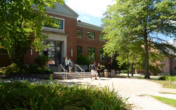 Brick building with steps and trees in sunlight. Students walk along a path, creating a lively, academic atmosphere on a clear day.