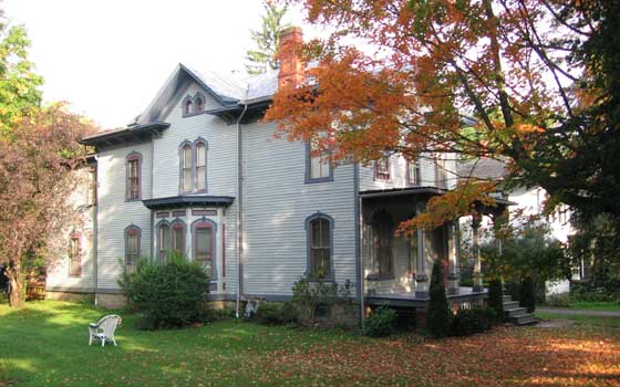 Victorian-style house with gray siding, tall windows, and mansard roof in autumn. Orange leaves cover the lawn; a white chair sits nearby.