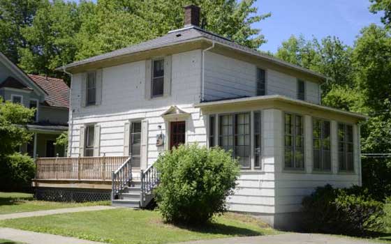 Two-story white house with a wooden porch, surrounded by lush greenery on a sunny day. Large windows and a small staircase lead to the entrance.