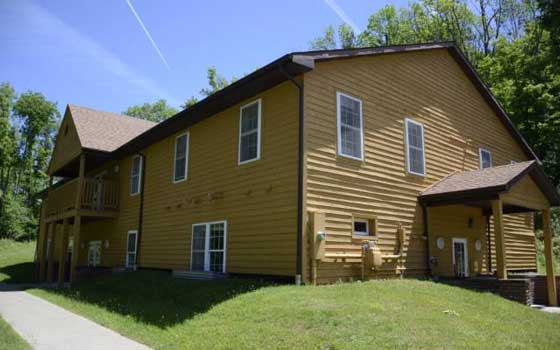 A two-story yellow house with a sloping roof stands amidst green grass and trees. The sun shines brightly, enhancing the vibrant summer atmosphere.