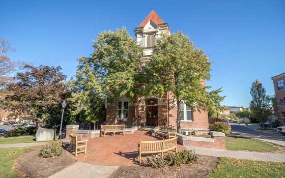 Historic brick building with large trees and benches in front, under a clear blue sky. Peaceful and inviting, with a classic architectural feel.