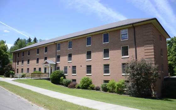 A three-story brick building with evenly spaced windows and a small entrance. Surrounded by green lawns, bushes, and trees under a clear blue sky.