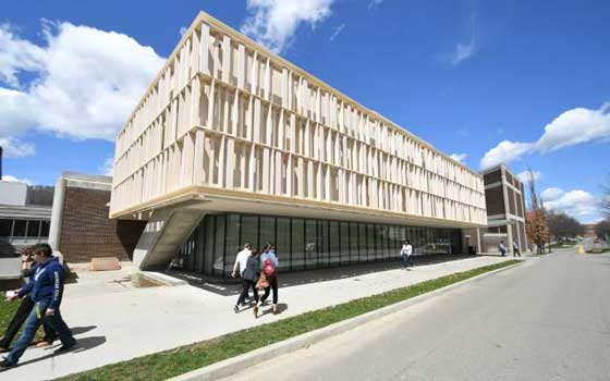 Modern building with unique wooden panel facade angles over a sidewalk. People walk below under a bright blue sky with a few clouds, conveying an academic setting.