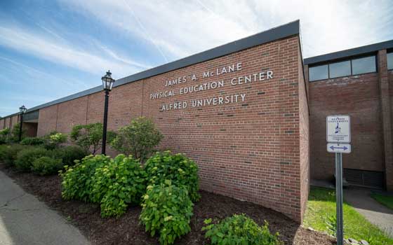 Red-brick building with "James A. McLane Physical Education Center, Alfred University" on the facade. Bushes line the walkway under a clear blue sky.