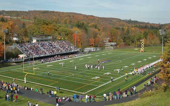 A football game in progress on a vibrant green field surrounded by trees with autumn foliage. A packed grandstand on the left, people watching, clear skies above.