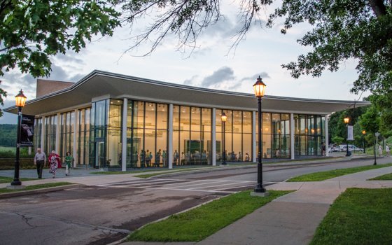 Modern building with large glass windows at dusk, emitting warm light. People are visible inside. Street lamps illuminate the sidewalk, creating a serene atmosphere.
