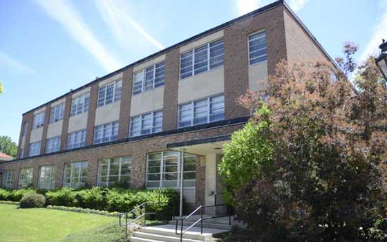 A three-story brick school building with large windows, surrounded by manicured bushes and a tree on a sunny day, conveying a peaceful academic setting.