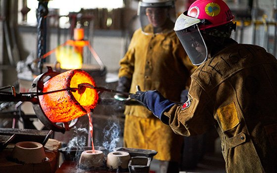 Two people in protective gear pour molten metal from a glowing crucible into molds. The scene shows intense heat and concentration.