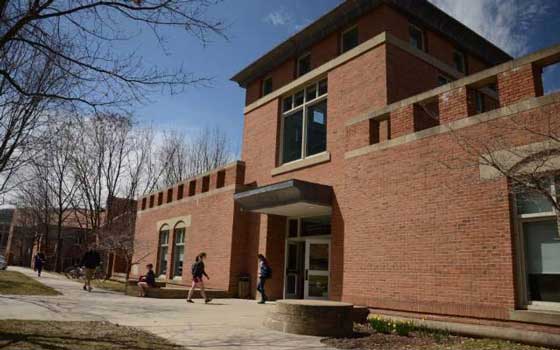 A modern brick college building under a clear blue sky, with students walking on the pathway. Leafless trees suggest late fall or early spring.