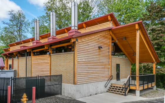 A modern wooden building with a red metal roof, four tall chimneys, and large overhangs. It is surrounded by trees, creating a natural setting.