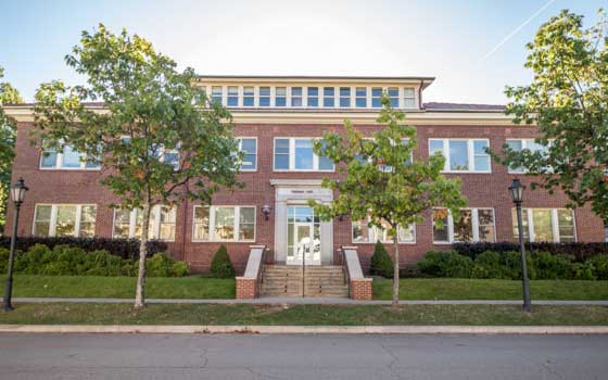 Red brick building with tall windows and a central staircase, flanked by trees and vintage lanterns. The scene conveys a calm, sunny ambiance.