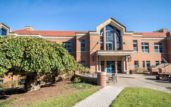 A brick building with large windows, a peaked entrance, and a patio with tables. A lush tree and green lawn are in the foreground under a clear blue sky.