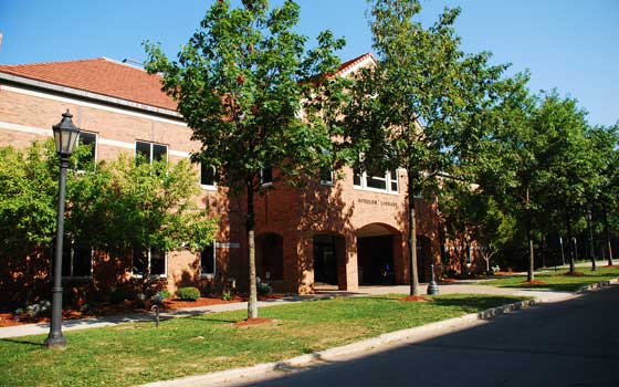 A brick building labeled "Scholes Library" is surrounded by leafy trees under a clear blue sky. The scene feels calm and inviting.
