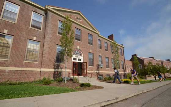 Three-story brick school building with large windows and a central entrance. People walk on the sidewalk in front. The tone is academic and inviting.