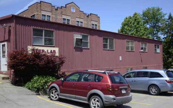 A maroon building with large windows and a "Lab" sign stands in a parking lot. A red car and a silver van are parked in front. The picture was taken in a bright and sunny setting.