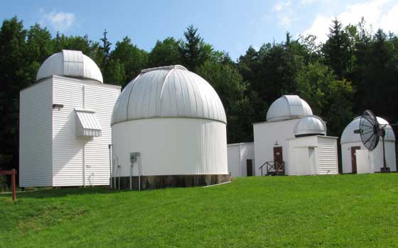Small observatory buildings with white domes set against a lush green lawn and dense trees. Clear blue sky suggests a peaceful, sunny day.