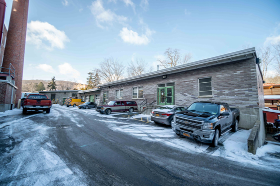 A snowy parking lot with several parked cars in front of a one-story brick building with a green door. Clear blue sky and leafless trees in the background evoke a cold, winter atmosphere.