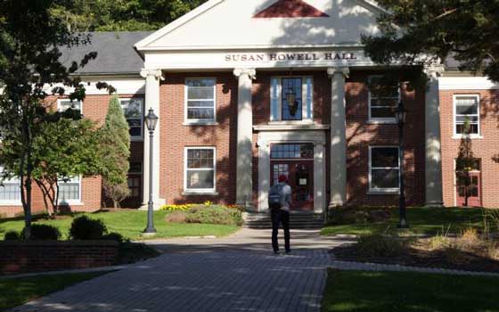 Brick building labeled "Susan Howell Hall" with white columns and green landscaping. A person is walking toward the entrance on a sunny day.